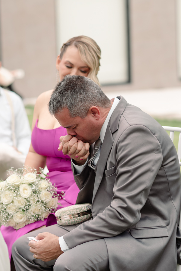Pai da noiva sentado na cerimônia em Jacareí, visivelmente emocionado, beijando as mãos enquanto segura um lenço. Fotografia de casamento na Chácara Mariah capturando a sensibilidade e o amor paterno durante o 
