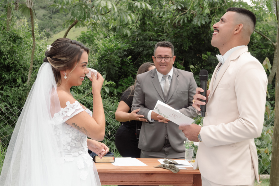 Noiva Larissa emocionada enxuga lágrimas enquanto o noivo Ricardo, visivelmente comovido e olhando para cima, lê seus votos durante a cerimônia ao ar livre. Fotografia profissional de casamento em Jacareí na Chácara Mariah capturando a pureza do momento.