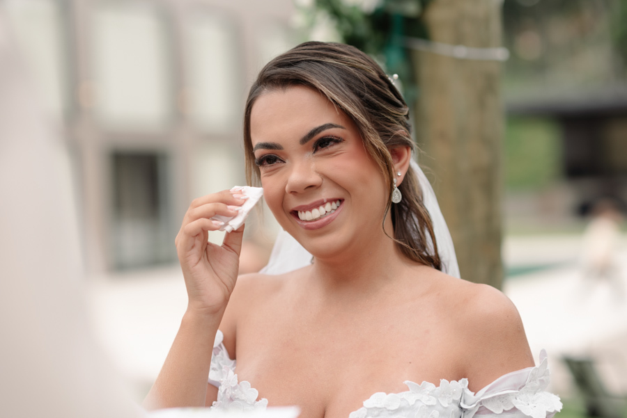 Noiva Larissa sorridente no altar em Jacareí, enxugando uma lágrima de felicidade com um lenço durante a cerimônia na Chácara Mariah. Fotografia de casamento profissional capturando a emoção e a beleza da noiva em seu momento especial.