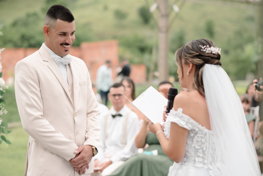 Noivo Ricardo sorri com ternura enquanto observa a noiva Larissa ler os votos durante a cerimônia ao ar livre. Fotografia profissional de casamento em Jacareí na Chácara Mariah, capturando a cumplicidade e o amor do casal no altar.