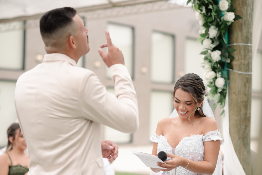Noiva Larissa sorri alegremente no altar enquanto o noivo Ricardo, de costas, aponta para o céu durante a leitura dos votos. Fotografia profissional de casamento em Jacareí na Chácara Mariah, capturando a espontaneidade e a fé do casal na cerimônia.
