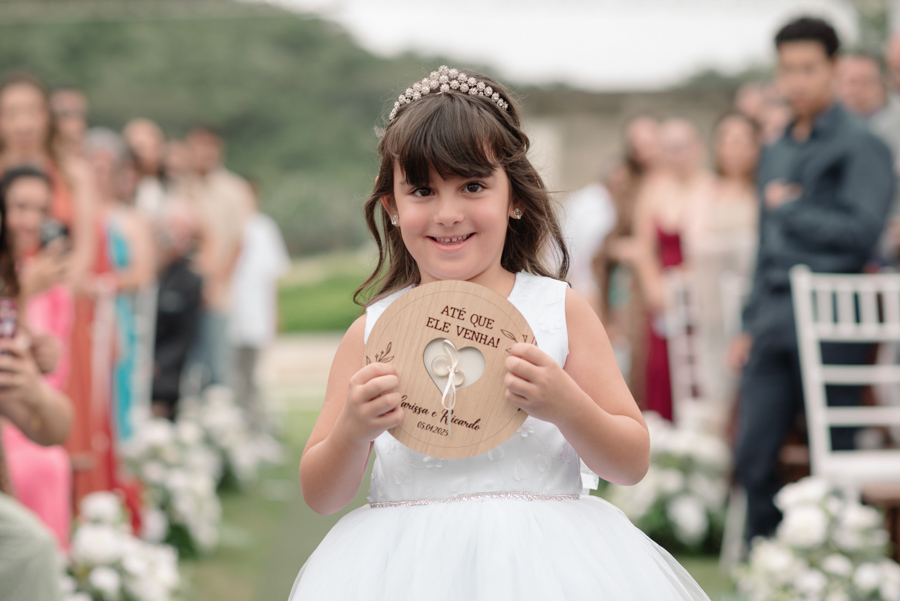 Daminha de honra sorridente em Jacareí, na Chácara Mariah, carregando as alianças em um suporte de madeira personalizado com a frase 