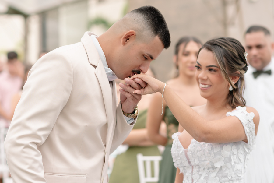Noivo Ricardo beija a mão da noiva Larissa após a troca de alianças na Chácara Mariah. Fotografia de casamento em Jacareí capturando o sorriso radiante da noiva e a reverência do noivo em momento de carinho e compromisso durante a cerimônia ao ar livre.