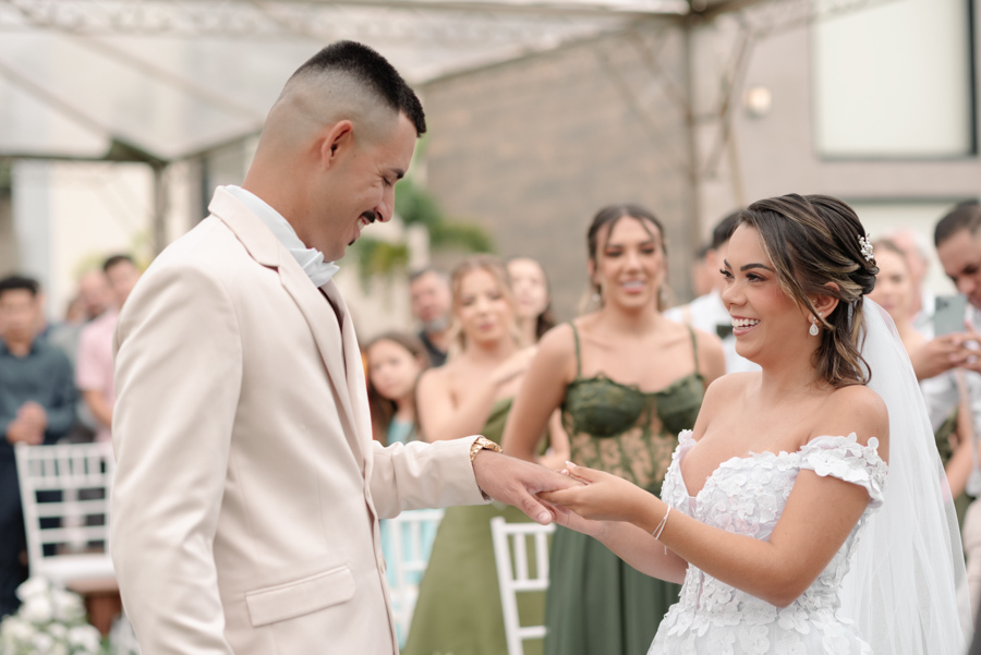 Noiva Larissa coloca a aliança no dedo do noivo Ricardo durante a cerimônia na Chácara Mariah. Fotografia de casamento em Jacareí capturando o casal sorridente e emocionado no altar, com as madrinhas ao fundo testemunhando o compromisso oficial.