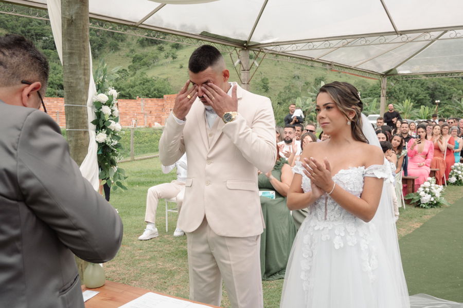 Noivo Ricardo enxuga lágrimas de emoção no altar ao lado da noiva Larissa, que aplaude durante a cerimônia na Chácara Mariah. Fotografia de casamento em Jacareí capturando a sensibilidade e alegria do casal após o 