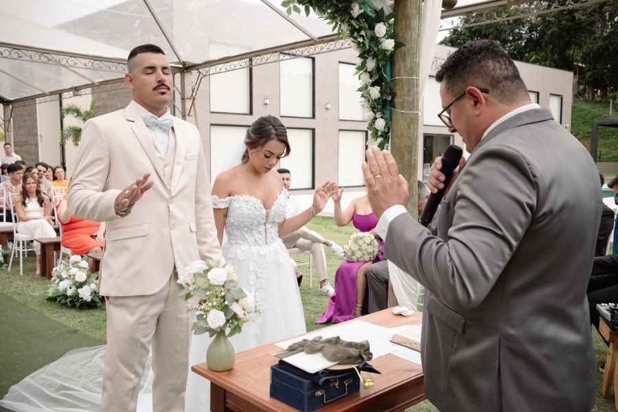 Noiva Larissa e noivo Ricardo de olhos fechados no altar enquanto o celebrante estende a mão em oração durante a cerimônia na Chácara Mariah. Fotografia de casamento em Jacareí capturando momento de fé e espiritualidade em celebração ao ar livre.