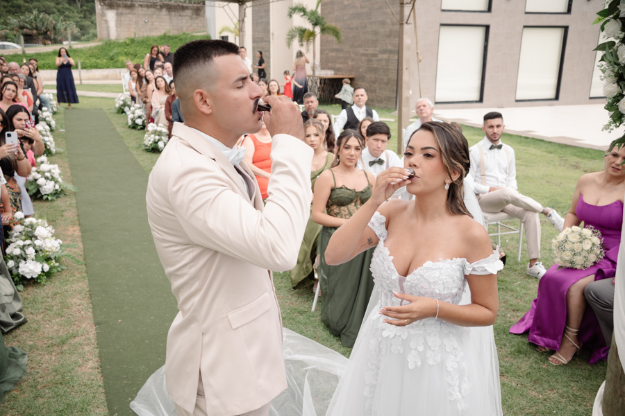 Noiva Larissa e noivo Ricardo participam do rito da Santa Ceia durante cerimônia ao ar livre na Chácara Mariah, em Jacareí. Fotografia de casamento capturando o casal bebendo o vinho no altar, simbolizando comunhão e fé diante dos convidados.