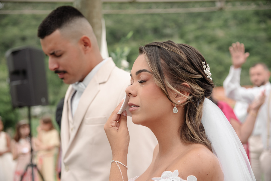 Noiva Larissa, visivelmente emocionada, enxuga as lágrimas com um lenço durante a cerimônia na Chácara Mariah, em Jacareí. Fotografia de casamento capturando a sensibilidade do casal no altar sob a luz natural, com o noivo Ricardo ao fundo.