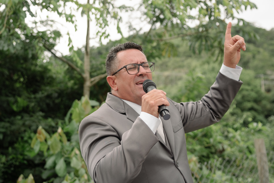 Celebrante de casamento em Jacareí canta com os olhos fechados e o dedo indicador apontado para o céu na Chácara Mariah. Fotografia profissional capturando a fé, a entrega e a espiritualidade durante a cerimônia cristã ao ar livre no Vale do Paraíba.