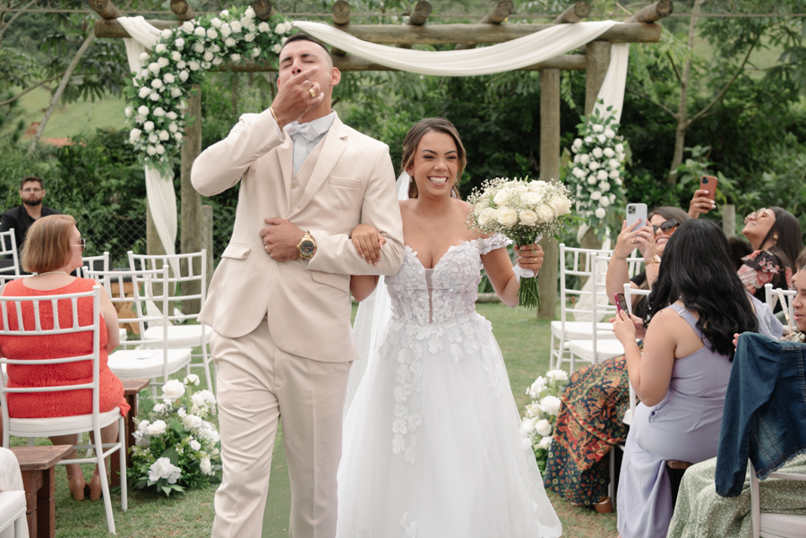 Noiva Larissa e noivo Ricardo saem do altar radiantes na Chácara Mariah, em Jacareí. O noivo manda um beijo para os convidados enquanto a noiva sorri com seu buquê de flores brancas, celebrando a união em um corredor de aplausos ao ar livre.