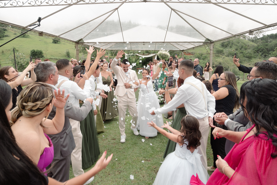 Noivos Larissa e Ricardo saem radiantes do altar sob chuva de pétalas brancas na Chácara Mariah, em Jacareí. Fotografia de casamento capturando a celebração dos convidados e a felicidade do casal em meio ao cenário campestre do Vale do Paraíba.