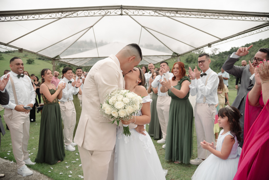 Noivos Larissa e Ricardo se beijam apaixonadamente no corredor após a cerimônia na Chácara Mariah, em Jacareí. Fotografia de casamento capturando a celebração dos convidados e madrinhas ao redor do casal em um cenário campestre sob a tenda aberta.