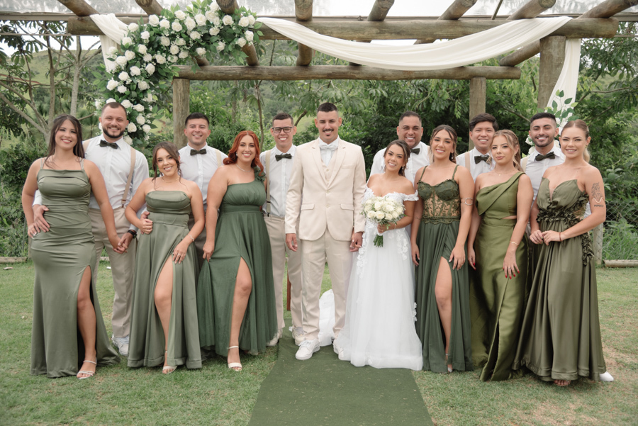 Noiva Larissa e noivo Ricardo posam no altar com seus padrinhos e madrinhas na Chácara Mariah, em Jacareí. Fotografia profissional destacando as madrinhas em diferentes tons de verde oliva e os padrinhos com suspensórios, em um cenário campestre ao ar liv