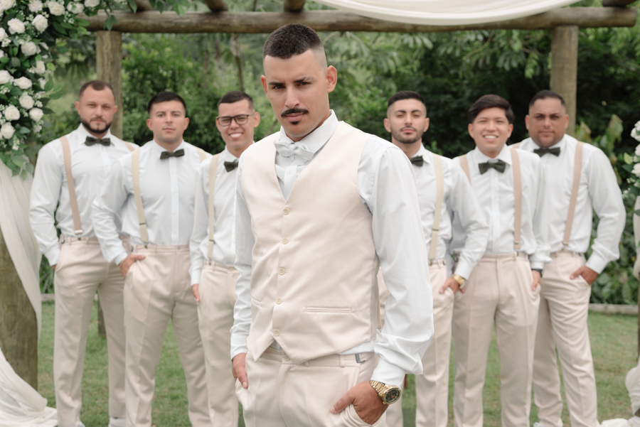 Noivo Ricardo posa em destaque à frente de seus padrinhos na Chácara Mariah, em Jacareí. Fotografia profissional de casamento capturando o noivo com colete bege e os padrinhos ao fundo, vestindo camisas brancas, suspensórios e gravatas borboleta verdes.