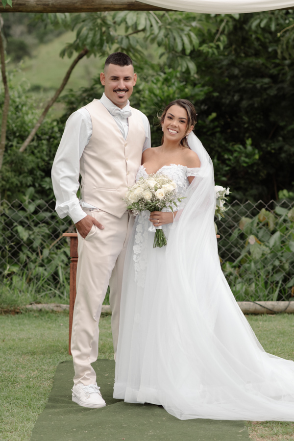Noiva Larissa e noivo Ricardo posam sorridentes no altar da Chácara Mariah, em Jacareí. Fotografia profissional de casamento capturando o casal de corpo inteiro; ele de terno bege e ela com vestido floral e longo véu branco em cenário campestre.
