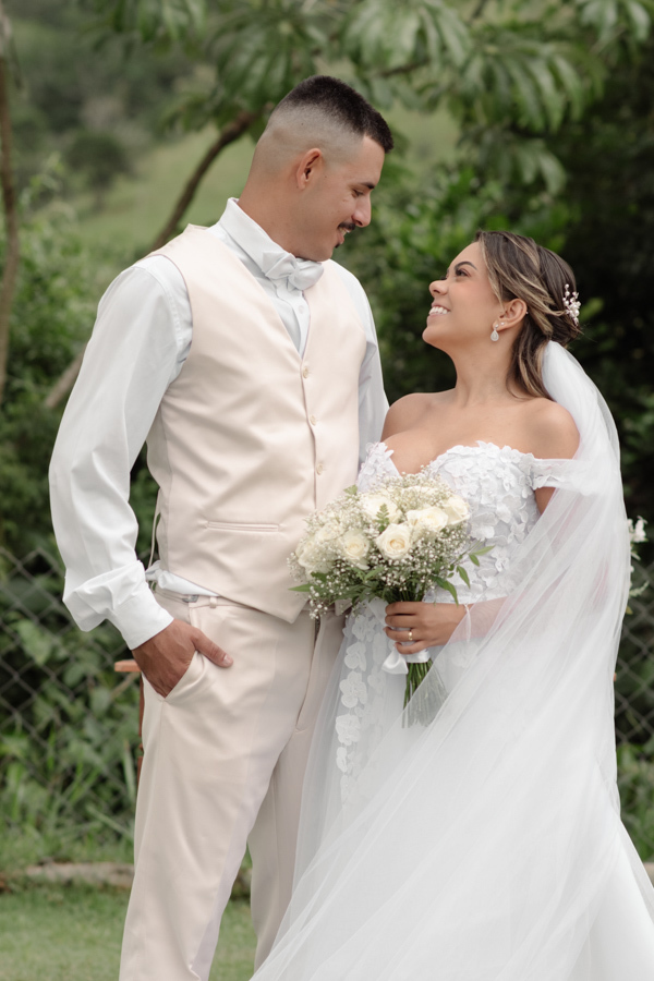 Noiva Larissa e noivo Ricardo posam sorridentes e trocam olhares no altar da Chácara Mariah, em Jacareí. Fotografia profissional de casamento capturando o casal de perfil; ele de colete bege e ela com vestido floral e longo véu branco em cenário campestre