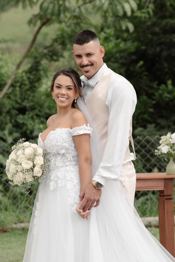 Noiva Larissa e noivo Ricardo posam sorridentes no altar da Chácara Mariah, em Jacareí. Fotografia profissional de casamento capturando o casal abraçado de lado; ela segura o buquê de flores brancas e ele veste colete bege em cenário campestre ao ar livre
