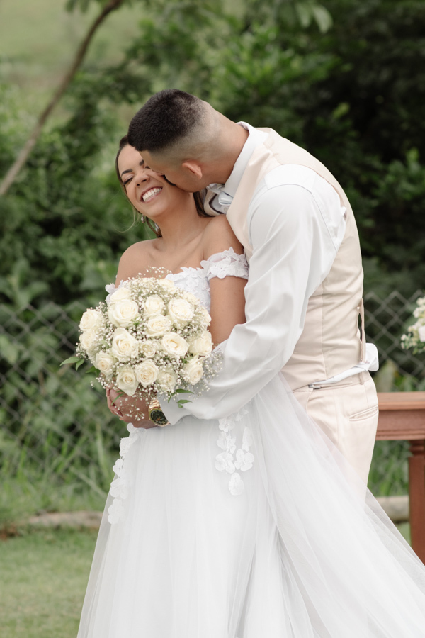 Noiva Larissa sorri alegremente enquanto o noivo Ricardo a abraça por trás e beija seu rosto durante ensaio na Chácara Mariah, em Jacareí. Fotografia profissional de casamento capturando momento de carinho e espontaneidade do casal em cenário campestre.