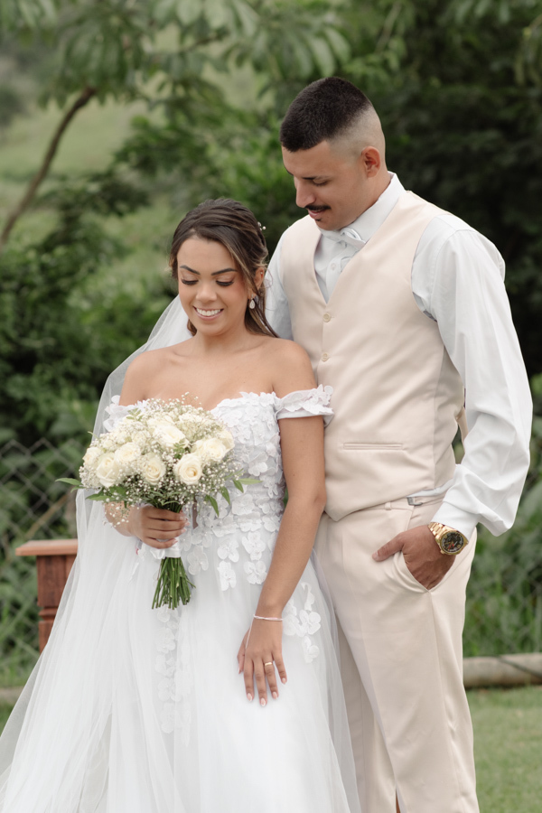 Noiva Larissa e noivo Ricardo posam para fotografia profissional na Chácara Mariah, em Jacareí. O casal está abraçado, com a noiva à frente segurando o buquê de rosas brancas e o noivo, vestindo colete bege, olhando-a com ternura em cenário campestre.