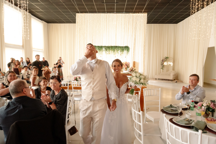 Noiva Larissa e noivo Ricardo entram sorridentes no salão de festas da Chácara Mariah, em Jacareí. Fotografia profissional capturando o casal sendo recebido com aplausos pelos convidados em ambiente decorado com luzes, celebrando o início da recepção.