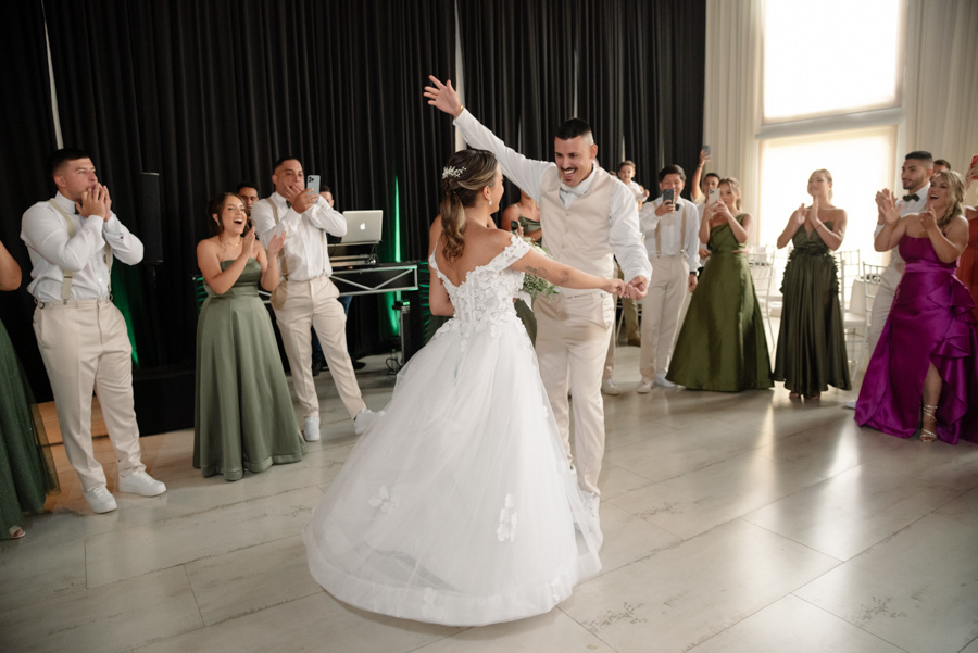 Noiva Larissa e noivo Ricardo dançam e celebram com animação no salão de festas da Chácara Mariah, em Jacareí. Fotografia de casamento capturando o casal no centro da pista, cercado por padrinhos e convidados que aplaudem e registram o momento com celular