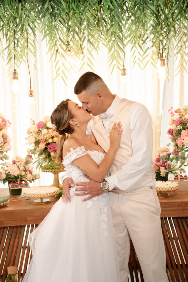 Noiva Larissa e noivo Ricardo se beijam apaixonadamente em frente à mesa de doces na recepção da Chácara Mariah, em Jacareí. Fotografia de casamento capturando momento romântico do casal sob decoração elegante com folhagens, flores e luzes pendentes.