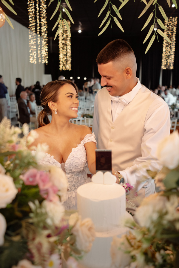 Noiva Larissa e noivo Ricardo sorriem e trocam olhares carinhosos ao cortarem o bolo na recepção da Chácara Mariah, em Jacareí. Fotografia de casamento capturando o casal à mesa decorada com flores, sob arranjos suspensos de folhagens e luzes decorativas.