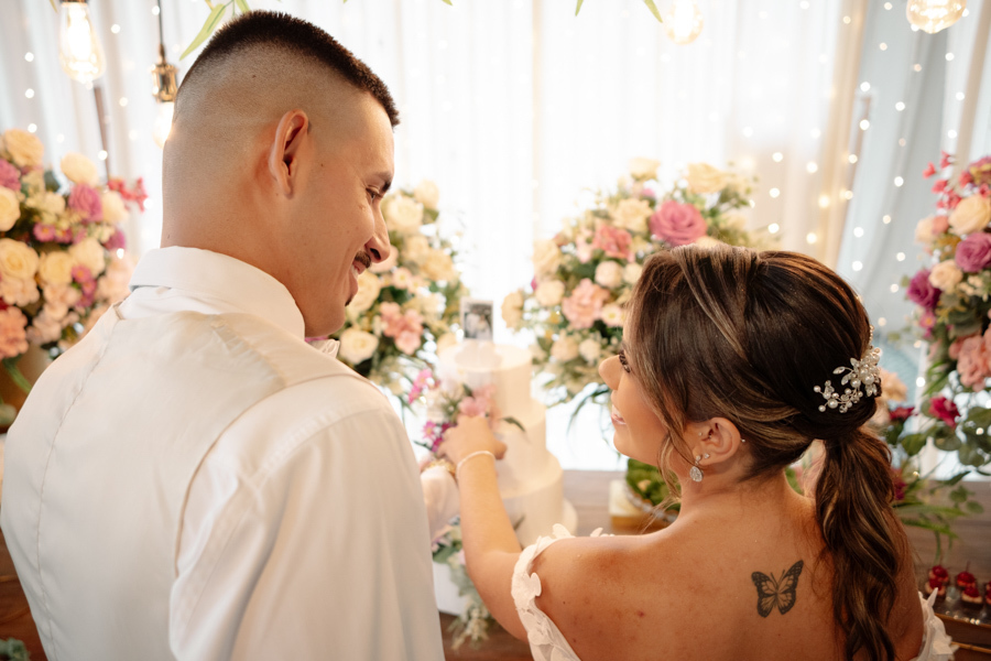 Noiva Larissa e noivo Ricardo sorriem e trocam olhares carinhosos ao cortarem o bolo na recepção da Chácara Mariah, em Jacareí. Fotografia de casamento capturando a noiva de costas, destacando sua tatuagem de borboleta e o penteado com arranjo de pérolas.