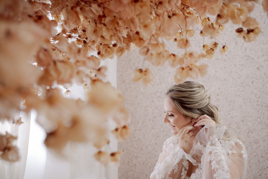 Noiva Claudia sorri ao colocar os brincos, vista através de flores secas decorativas. Fotografia artística de making of de casamento em Jacareí, capturando a delicadeza e os preparativos finais pelo fotógrafo Gilson Dias.