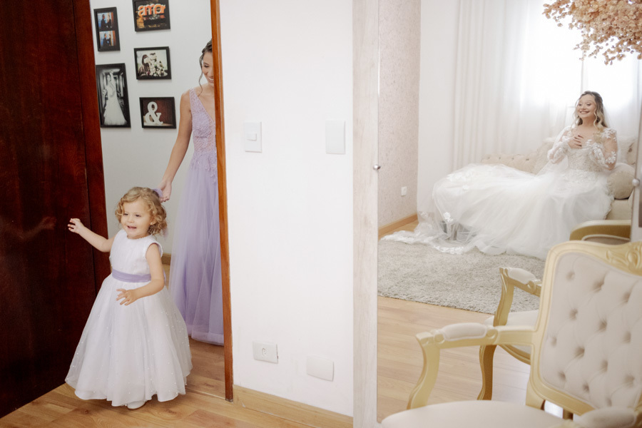 Daminha de honra entrando no quarto e surpreendendo a noiva Claudia durante o making of em Jacareí. Registro espontâneo de emoção e alegria refletida no espelho, capturado pelo fotógrafo Gilson Dias em um momento único do casamento.