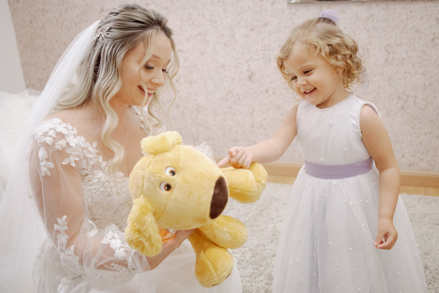 Noiva Claudia e daminha de honra sorriem juntas brincando com mascote de pelúcia durante o making of em Jacareí. Fotografia espontânea de casamento capturando a pureza e alegria do momento por Gilson Dias.