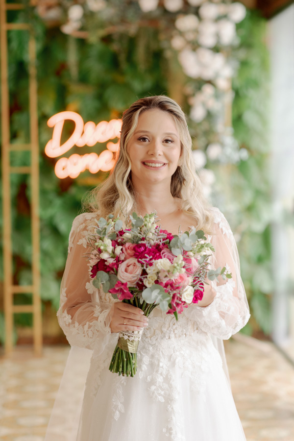 Retrato da noiva Claudia sorridente segurando buquê de flores rosa e lilás. Ao fundo, painel de folhagens com letreiro neon decorativo. Fotografia de casamento em Jacareí capturando a elegância da noiva pelo fotógrafo Gilson Dias.