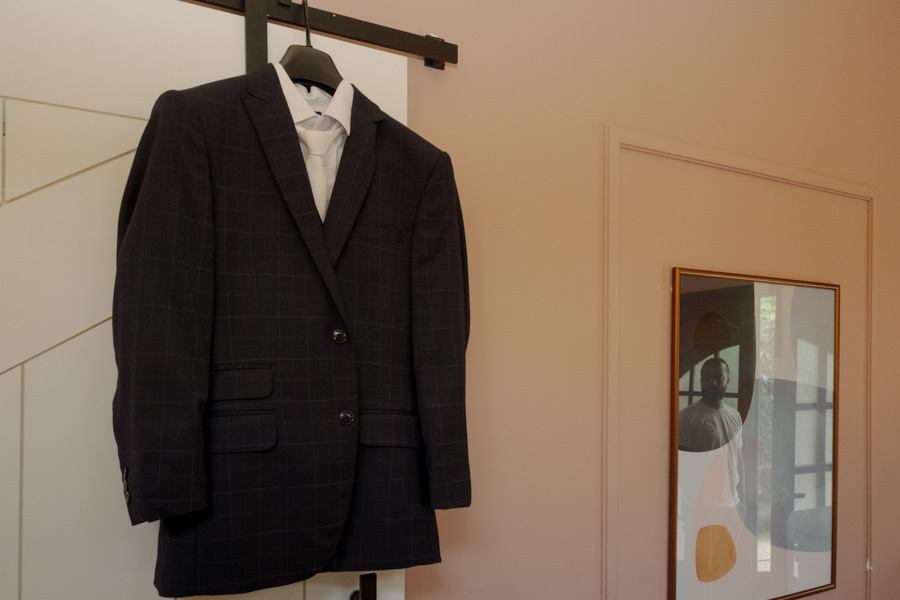 Traje do noivo Matheus pendurado durante o making of em Jacareí. Detalhe do terno azul marinho com gravata clara, preparando o cenário para a fotografia de casamento capturada pelo olhar atento de Gilson Dias.