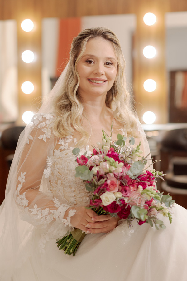 Retrato da noiva Claudia sentada com seu buquê de flores em tons de rosa, destacada por luzes de camarim ao fundo. Fotografia de noiva em Jacareí capturando a elegância e o brilho do making of pelo fotógrafo Gilson Dias.