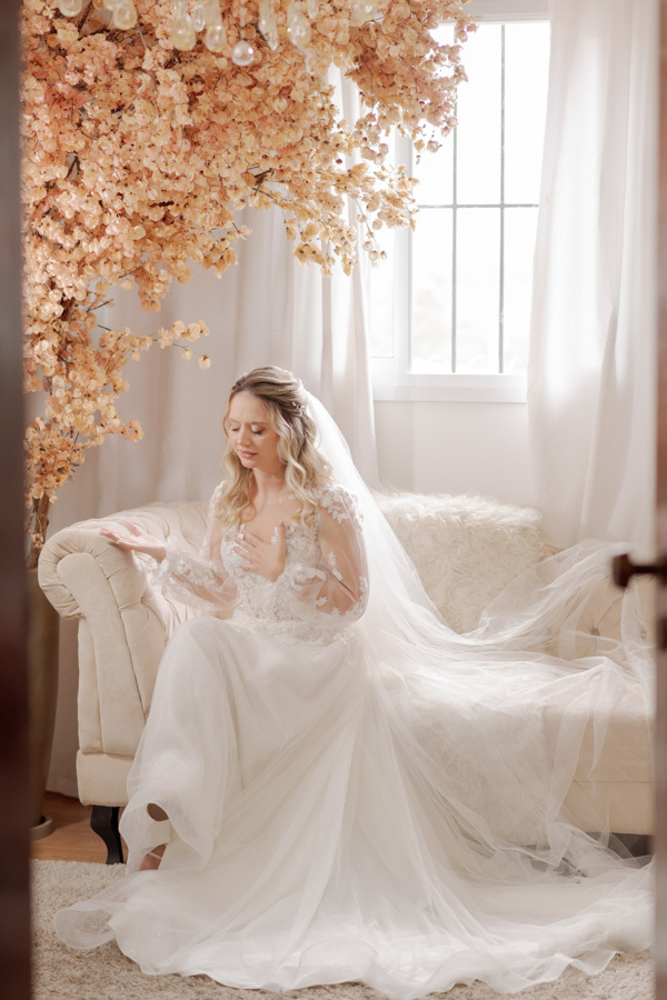 Noiva Claudia em momento de reflexão e oração sentada no sofá durante o making of em Jacareí. O véu longo se estende pelo cenário iluminado com decoração floral, capturado com sensibilidade pelo fotógrafo Gilson Dias.