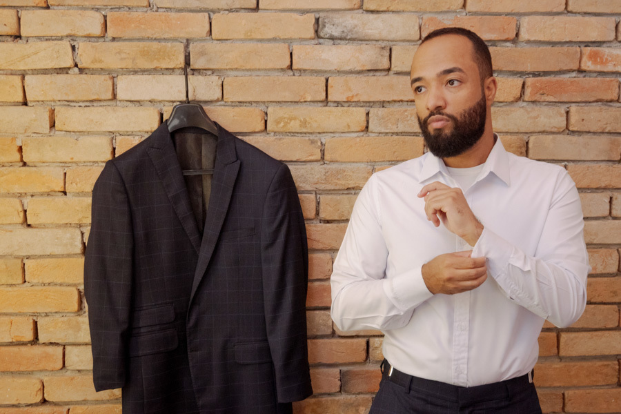 Making of do noivo Matheus em Jacareí: noivo ajusta os punhos da camisa branca ao lado de seu terno azul pendurado em parede de tijolos aparentes. Fotografia de casamento com olhar documental e autêntico capturada por Gilson Dias.