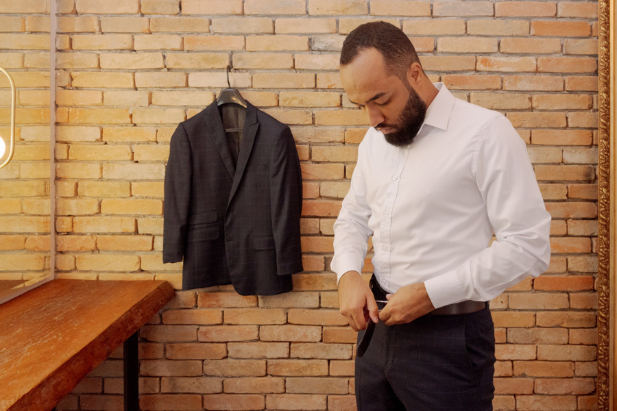 Making of do noivo Matheus em Jacareí: noivo ajusta o cinto do traje social ao lado de seu terno azul pendurado em parede de tijolos. Fotografia de casamento focada nos preparativos e detalhes masculinos, capturada pelo fotógrafo Gilson Dias.