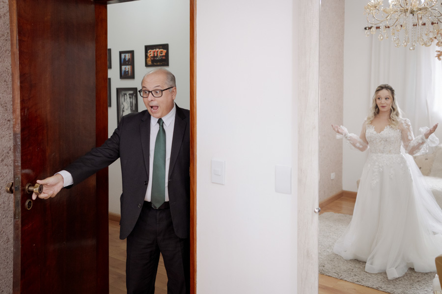 Daminha de honra entrando no quarto e surpreendendo a noiva Claudia no making of em Jacareí. Registro espontâneo de alegria refletida no espelho, capturando a emoção real do casamento pelo fotógrafo Gilson Dias.