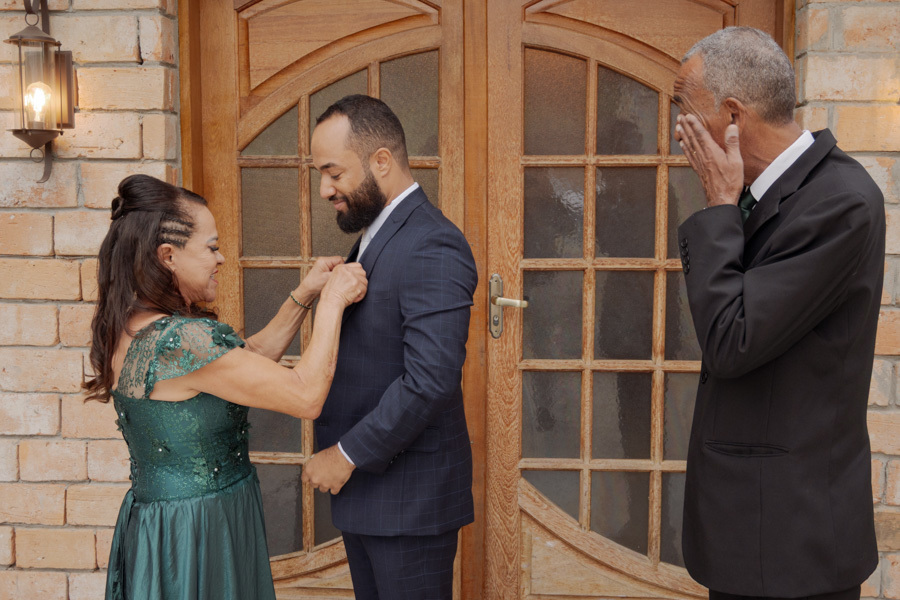 Making of do noivo Matheus em Jacareí: mãe ajusta o terno do filho enquanto o pai se emociona ao fundo. Registro tocante de cumplicidade familiar em casamento no Boulevard, capturado com sensibilidade pelo fotógrafo Gilson Dias.