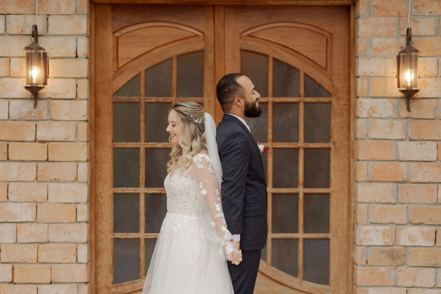 First touch emocionante: noiva Claudia e noivo Matheus de costas um para o outro, de mãos dadas em frente a uma porta rústica de madeira em Jacareí. Fotografia de casamento capturando a ansiedade e o amor antes do altar por Gilson Dias.