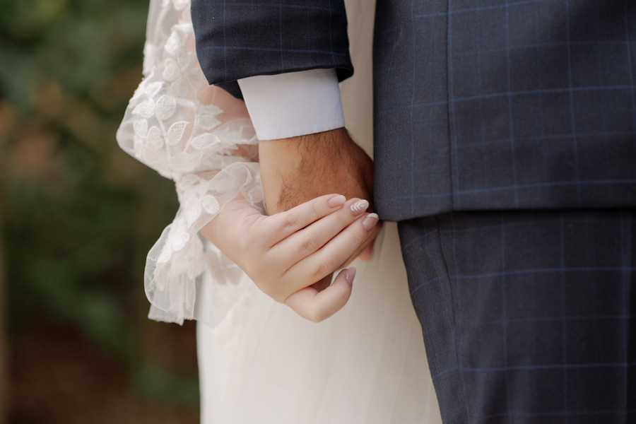 Close detalhado das mãos dadas da noiva Claudia e do noivo Matheus durante o first touch em Jacareí. Destaque para a delicadeza da renda do vestido e o terno do noivo, capturando a conexão do casal pelo fotógrafo Gilson Dias.