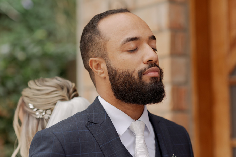 Close do noivo Matheus de olhos fechados durante o first touch em Jacareí. Ao fundo, a noiva Claudia posicionada de costas. Fotografia de casamento que captura a emoção, oração e conexão do casal antes da cerimônia por Gilson Dias.