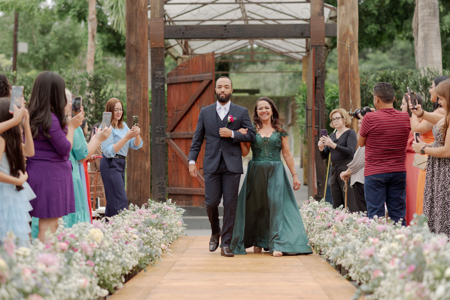 Entrada do noivo Matheus com sua mãe em cerimônia ao ar livre no Boulevard Jacareí. Corredor decorado com flores, convidados registrando o momento e fotografia de casamento capturando a emoção da caminhada ao altar por Gilson Dias.