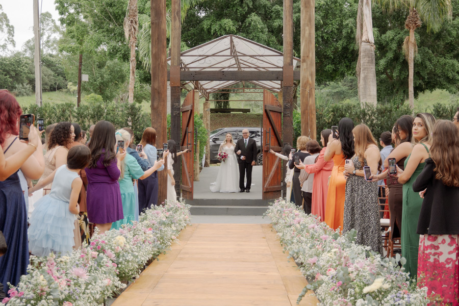 Entrada da noiva Claudia com seu pai em cerimônia ao ar livre no Boulevard Jacareí. Convidados emocionados registram a caminhada pelo corredor decorado com flores. Fotografia de casamento espontânea e marcante por Gilson Dias.