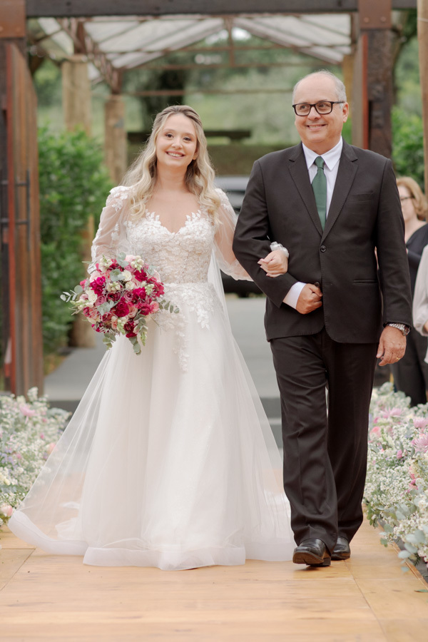 Noiva Claudia caminha sorridente com seu pai pelo corredor da cerimônia ao ar livre no Boulevard Jacareí. Destaque para o vestido de renda e o buquê rosa em um momento de pura felicidade registrado pelo fotógrafo Gilson Dias.