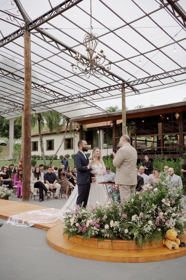 Cerimônia de casamento de Claudia e Matheus no Boulevard Jacarei. Vista ampla do altar decorado com flores e lustre rústico, capturando o casal diante do celebrante em um cenário ao ar livre. Fotografia documental por Gilson Dias.