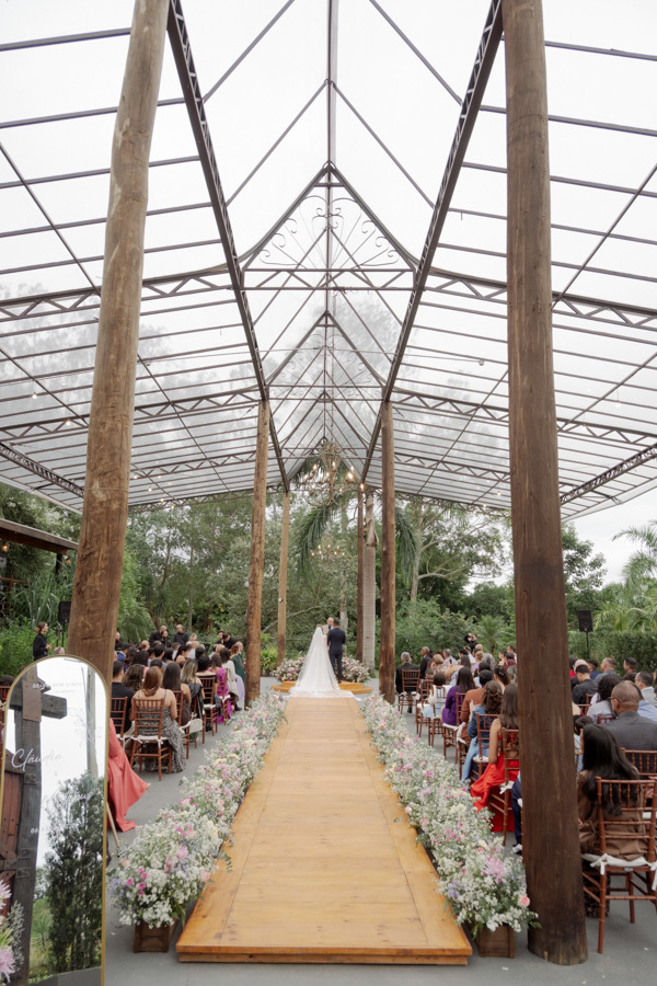 Vista panorâmica da cerimônia de Claudia e Matheus no Boulevard Jacareí. O corredor ladeado por flores leva ao altar sob uma estrutura rústica e imponente. Fotografia de casamento que captura a grandiosidade do evento por Gilson Dias.
