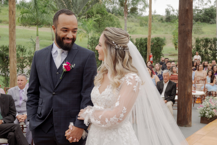Noivo Matheus e noiva Claudia trocam olhares e sorrisos de mãos dadas no altar em Jacareí. Fotografia de casamento no Boulevard capturando a cumplicidade e o amor do casal durante a cerimônia ao ar livre, registrada por Gilson Dias.