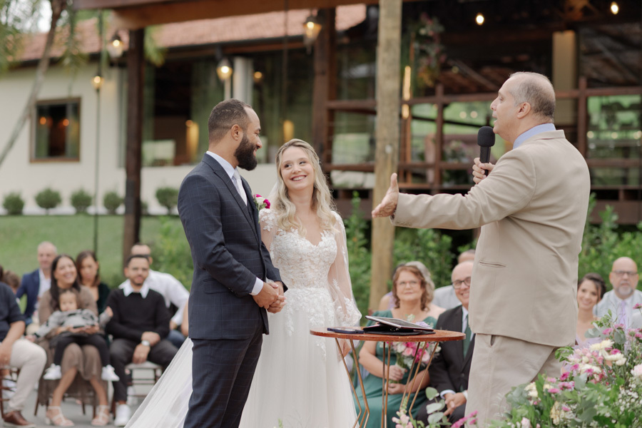 Noivos Claudia e Matheus sorriem para o celebrante no altar do Boulevard Jacareí. Fotografia de casamento capturando a alegria do casal e a atenção dos convidados em cerimônia ao ar livre, registrada pelo fotógrafo Gilson Dias.