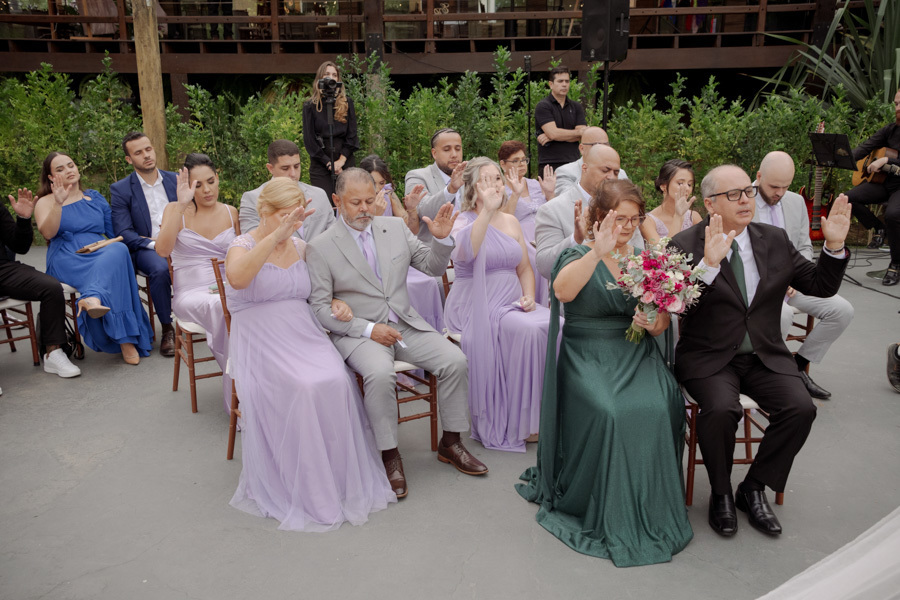 Convidados e padrinhos estendem as mãos em benção durante a cerimônia de Claudia e Matheus no Boulevard Jacareí. Fotografia de casamento capturando um momento de fé e união ao ar livre, registrada pelo fotógrafo Gilson Dias.